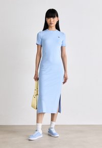 Femme se tenant debout, portant une robe midi ajustée bleu clair avec une fente sur le côté, des baskets assorties, des chaussettes blanches et tenant un sac à main jaune pâle.