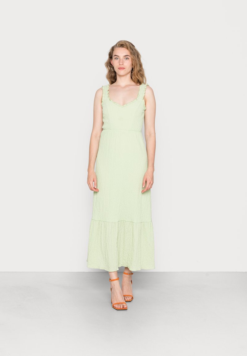 Vestido maxi verde claro com alças frisadas, tecido texturizado e bainha em camadas. Combinado com sandálias de salto alto laranja com tiras.