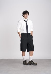 Young man standing with hands in pockets, wearing white shirt with black tie, black long shorts, white socks, and black sneakers against plain background.