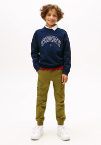 Mørkeblå sweatshirt med "TOMMY" trykk, hvit krage, olivenfargede cargo-bukser med lommer, og hvite joggesko med snørebånd. Avslappet barneantrekk.