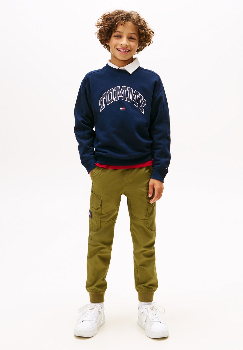Mørkeblå sweatshirt med "TOMMY" trykk, hvit krage, olivenfargede cargo-bukser med lommer, og hvite joggesko med snørebånd. Avslappet barneantrekk.