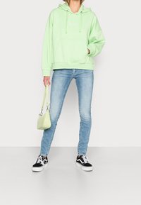 Felpa verde chiaro con tasca frontale e cordini, abbinata a jeans azzurri chiari. Sneakers nere e bianche e una piccola borsa verde.