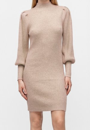 Beige ribstrikket kjole med høj hals og lange puffede ærmer, tætsiddende silhuet, der ender over knæet på en stående kvinde.
