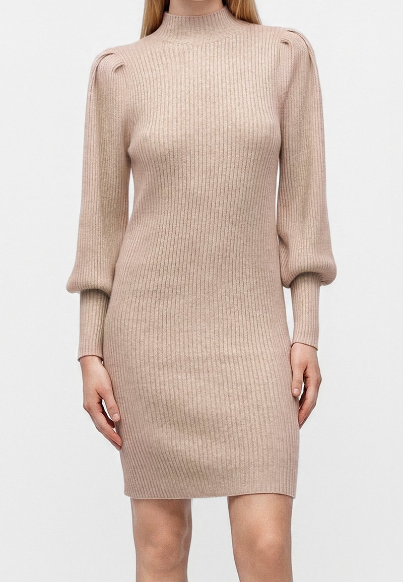Robe en maille côtelée beige avec col montant et manches longues bouffantes, silhouette ajustée se terminant au-dessus du genou portée par une femme debout.
