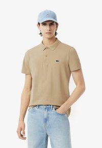 Camisa polo de algodón beige con cuello y tres botones, con un logo bordado en color azul oscuro. Combinada con unos pantalones vaqueros de mezclilla azul claro y una gorra azul claro.