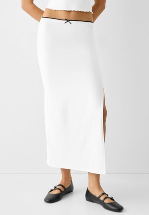 Biała długa spódnica maxi z bocznym rozcięciem, z czarnym paskiem w talii i kokardą. Noszona z czarnymi balerinami. Gładka faktura tkaniny.