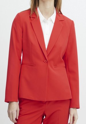 Vrouw draagt een nauwsluitend rood colbert met één knoop over een wit overhemd met kraag, gecombineerd met bijpassende rode broek.