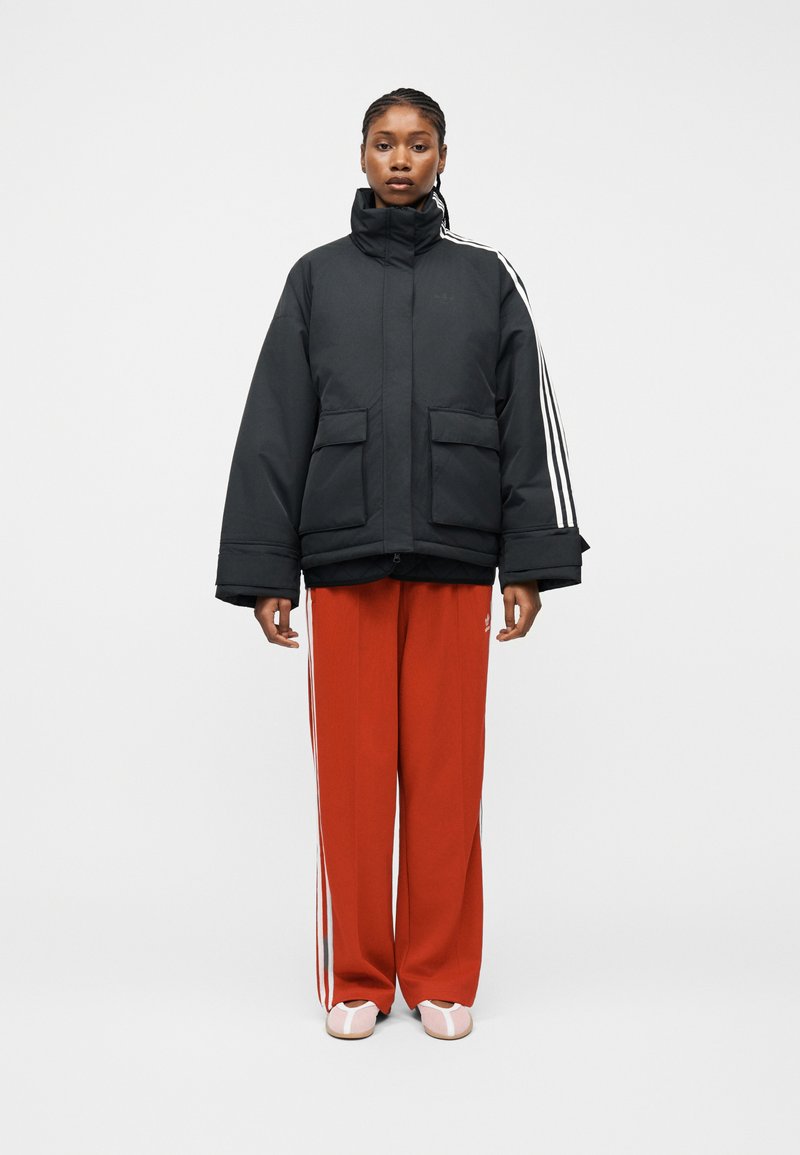 Γυναίκα που στέκεται προς τα εμπρός φορώντας μαύρο μπουφάν τύπου puffer με λευκές ρίγες, κόκκινο παντελόνι φόρμας με λευκές ρίγες και ροζ παπούτσια.