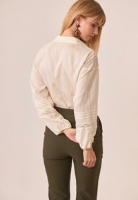 Blusa color crema de manga larga con detalles de encaje y puños en capas, combinada con pantalones ajustados de color verde oscuro.