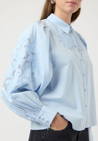 Ανοιχτό μπλε πουκάμισο με φουσκωτά μανίκια, λεπτομερές κεντητό λουλουδάτο σχέδιο στους ώμους και κουμπιά μπροστά. Διαθέτει μανσέτες με ρέλια και κλασικό γιακά.
