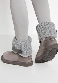 Pies que llevan botas UGG de ante gris con puños de punto doblados y pantalones ajustados en color beige, mostrando el patrón de la suela en una de ellas.
