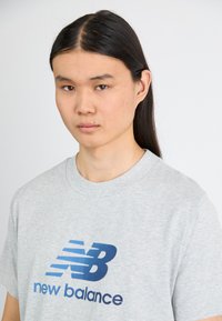 Gray short-sleeve T-shirt featuring a blue "New Balance" logo with diagonal lines, made of soft cotton fabric. Classic crew neck design.