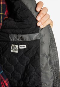 Hand die een open zwarte gewatteerde jas houdt, waarbij de binnenkant wordt getoond met een drukknopzak, een stoffen label met QR-code en een klein wit etiket.