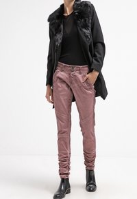 Svart kappa med fuskpälskrage, långärmad svart topp och rosa texturerade byxor med knapppryttare och skrumpna ärmslut. Svarta anklarstövlar.