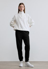 Άσπρο φούτερ με φερμουάρ και ριμπ μανσέτες, συνδυασμένο με μαύρο παντελόνι φόρμας με μικρό πράσινο λογότυπο και λευκά αθλητικά παπούτσια.