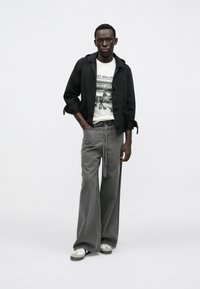 Čierna ľahká bunda s kapucňou, biele tričko s grafickým motívom a sivé široké nohavice. Tenisky majú biele a sivé detaily.