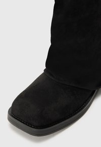 Botte en daim noir avec un bout carré et une semelle noire légèrement surélevée sur un fond gris clair.