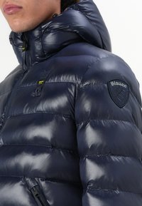 Marine blauwe pufferjack met een glanzende afwerking, voorzien van een capuchon, ritszakken en een ronde patchlogo op de schouder.