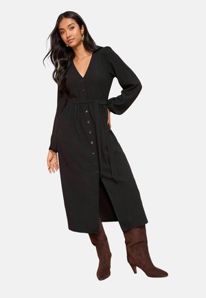 Must pika varrukaga kleit V-kaelusega, ees nööpide ja vöökohaga lipsuga. A-lõikeline siluett kõrge lõhikuga, sobitatud pruunide põlveni ulatuvate saapadega.