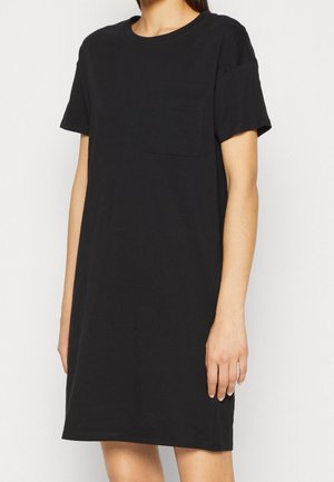 Kobieta ubrana w prostą czarną sukienkę typu T-shirt z krótkim rękawem i małą kieszonką na piersi, stojąca na białym tle.