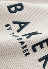 Primer plano de una tela texturada de color crema con grandes letras bordadas en negro "BAKER" y un texto más pequeño "BY TRABAHER".