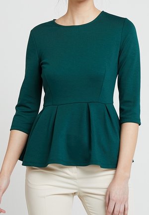 Kvinde iført en mørkegrøn, tætsiddende peplum-top med trekvartlange ærmer og lyse beige bukser mod en ensfarvet baggrund.