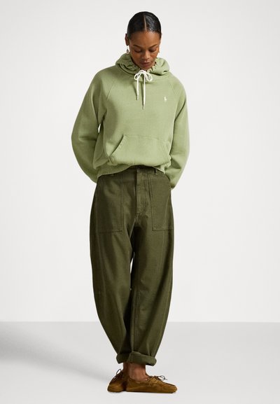 Camisola com capuz verde com cordão, bolso frontal tipo canguru, usada com calças largas verde-oliva. Sapatos castanhos estilo mocassim, modelo num cenário neutro.