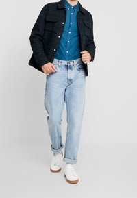 Conjunto masculino que incluye una chaqueta de cuadros oscura, camisa de botones color teal, vaqueros azul claro y zapatillas blancas con suelas marrones.