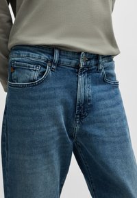 Blue denim jeans with a mid-rise fit, five pockets, and a zip fly. Features slight fading and stitching details. Textured fabric.