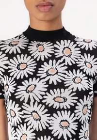 Nærbillede af en person iført en sort kortærmet top med et hvidt tusindfryd blomstermønster og ferskenfarvede blomstercentre.