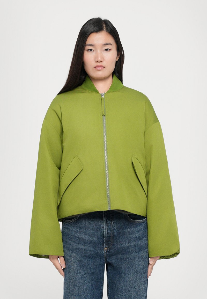 Green cropped bomber jacket with a stand-up collar, front zipper, and side pockets. Smooth texture and wide sleeves. Worn over blue jeans.