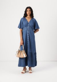 Denim maksi haljina s kratkim zvonastim rukavima, nabrani korzet, dugme sprijeda i višeslojni rub. Model drži malu torbicu u više boja.