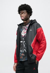 Αντιανεμικό μαύρο και κόκκινο με φερμουάρ, λευκές λεπτομέρειες και λογότυπο Jordan. Συνδυασμένο με γραφικό μπλουζάκι που απεικονίζει μπάσκετ.
