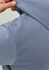 Jasnoniebieska bluza z kapturem wykonana z teksturowanej mieszanki bawełny, z kapturem na sznurku oraz zbliżenie na szew ramienia.