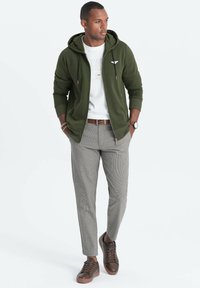 Sudadera verde oliva con cremallera sobre una camisa blanca, combinada con pantalones de houndstooth gris claro y zapatillas marrones. Los accesorios incluyen un reloj.