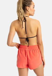 Korallenrosa Shorts mit elastischem Bund, Seitentaschen und glatter Textur, kombiniert mit einem schwarzen Neckholder-Oberteil, das im Rücken gebunden ist.