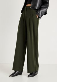 Άτομο που φοράει ψηλόμεσες φαρδιές ελαιόκαφε παντελόνες, μαύρη μπουφάν δερματίνης, και μαύρες γόβες με τακούνι, που στέκεται σε λευκό δάπεδο.