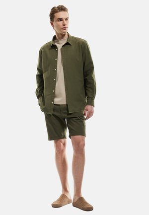 Oliven grøn, knap-op skjorte over en beige t-shirt, parret med matchende oliven grønne shorts og lysebrune slip-on sko, stående stilling.