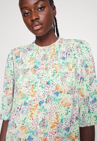 Blusa floral de botões com mangas bufantes. Apresenta um tecido verde pastel com padrões de flores multicoloridas e uma textura subtil. Gola redonda.