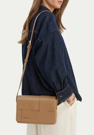 Mujer con chaqueta de mezclilla azul oscuro y pantalones blancos, llevando un bolso cruzado de cuero marrón claro con una correa larga ajustable.