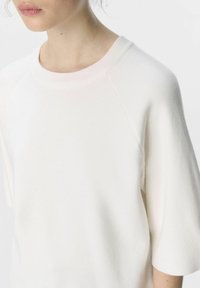 Pull blanc avec un col rond à côtes, des manches raglan courtes et une texture douce et lisse. Le tissu semble léger et douillet.