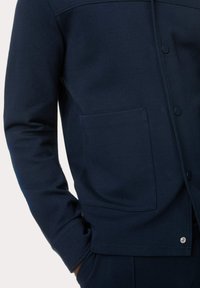 Giacca button-up blu navy con tessuto morbido, grande tasca frontale e dettagli di cucitura discreti lungo le spalle.
