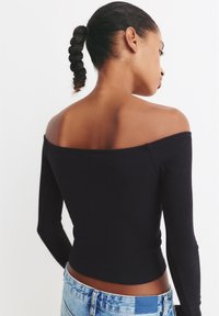 Schwarzes, geripptes Langarmoberteil mit schulterfreiem Ausschnitt. Figurbetontes Design, leicht tailliert, getragen mit ausgebleichten blauen Jeans.