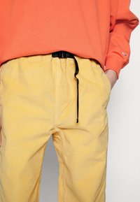 Jasnożółte spodnie dżinsowe z teksturowanej tkaniny, z elastycznym pasem i czarnym sznurkiem, w zestawie z pomarańczową bluzą.