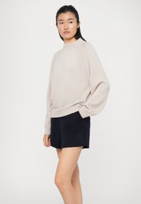 Maglione oversize beige chiaro con collo alto e maniche raglan, abbinato a pantaloni corti blu navy con tasche. Texture liscia.