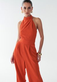 Oranžový overal na ramínka s textúrovanou tkaninou, skladaným dizajnom a širokými nohami. Doplnok v podobe zodpovedajúceho pásu a zlatých šperkov.
