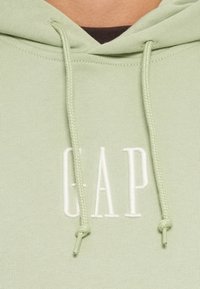 Felpa verde chiaro con cappuccio e cordino, con la scritta "GAP" in lettere bianche ricamate. Tessuto liscio, spessore medio.