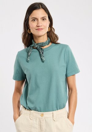 Femme aux cheveux bruns ondulés portant un t-shirt manches courtes bleu sarcelle, une écharpe à motifs verts autour du cou, des boucles d'oreilles anneaux dorés, et un pantalon beige avec les mains dans les poches.