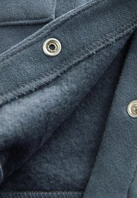 Tejido azul oscuro con forro suave, que presenta cierres a presión de metal y costuras visibles. Muestra una variación de textura entre los materiales exterior e interior.