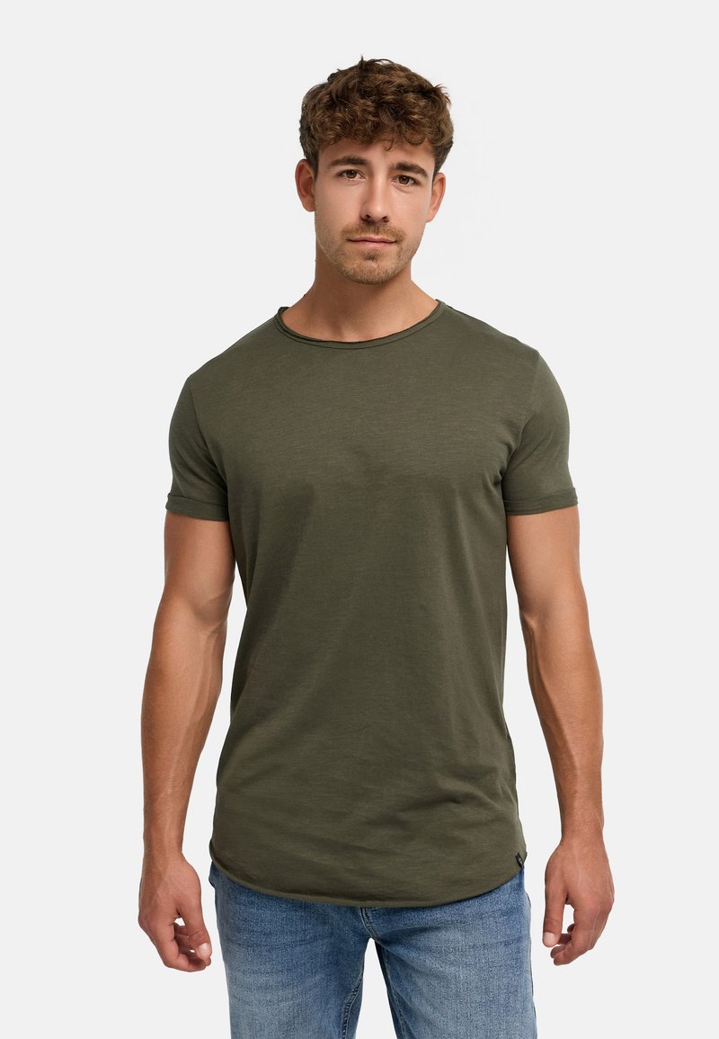 Jeune homme aux cheveux courts et bouclés portant un t-shirt manches courtes vert olive et un jean bleu sur un fond clair uni.